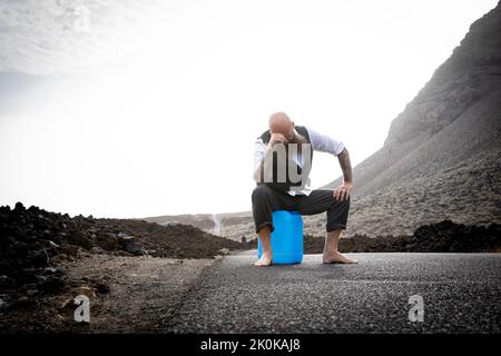 L'uomo con vestito è seduto completamente disperato e bussato fuori su un canister blu da qualche parte in nessun posto nel deserto vulcanico alla fine o Foto Stock