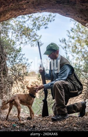 Vista laterale del cacciatore maschio in mimetizzazione e cappello in piedi sul ginocchio mentre accarezzano il cane da caccia tra le rocce in campagna Foto Stock