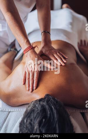 Taglia irriconoscibile massaggiatore femminile in uniforme bianca facendo massaggio alla schiena per l'uomo sdraiato sul divano in sala salone luminoso di giorno Foto Stock