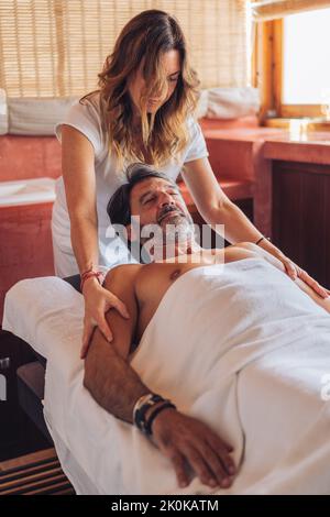 Donna in bianco uniforme massaggiante spalle di contenuti uomo maturo sdraiato sul tavolo sul retro con gli occhi chiusi in accogliente salone Foto Stock