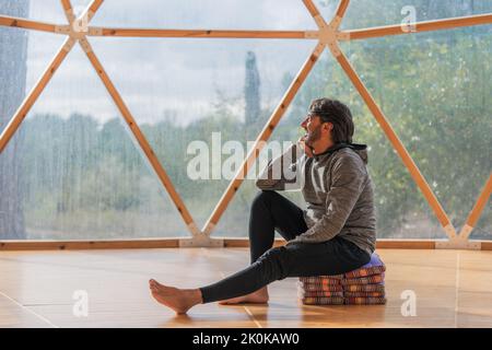 Vista laterale di un allegro uomo anziano bearded grigio capelli con piedi nudi e mento che riposa a mano seduto su coperte piegate in una stanza di legno vicino panoramico Foto Stock