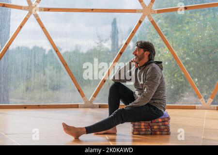 Vista laterale di un allegro uomo anziano bearded grigio capelli con piedi nudi e mento che riposa a mano seduto su coperte piegate in una stanza di legno vicino panoramico Foto Stock