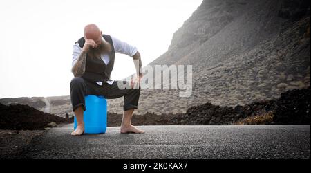 L'uomo con vestito è seduto completamente disperato e bussato fuori su un canister blu da qualche parte in nessun posto nel deserto vulcanico alla fine o Foto Stock