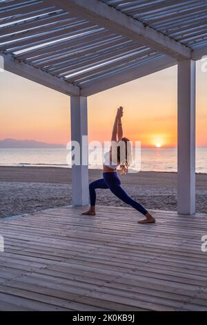 Vista laterale della tranquilla donna in abbigliamento sportivo che pratica yoga in Crescent Lunge posa mentre si trova in piedi sulla terrazza di legno vicino al mare durante l'alba Foto Stock