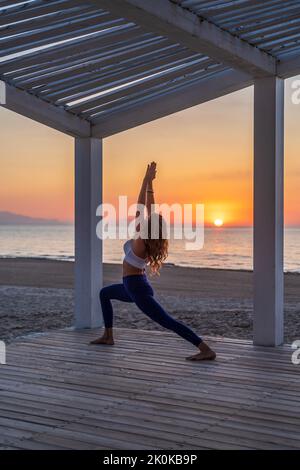 Vista laterale della tranquilla donna in abbigliamento sportivo che pratica yoga in Crescent Lunge posa mentre si trova in piedi sulla terrazza di legno vicino al mare durante l'alba Foto Stock