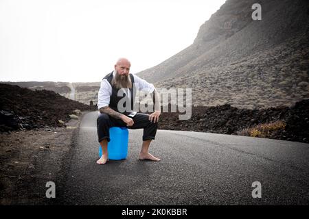 L'uomo con vestito è seduto completamente disperato e bussato fuori su un canister blu da qualche parte in nessun posto nel deserto vulcanico alla fine o Foto Stock