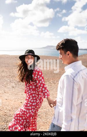 Vista laterale di una giovane coppia etnica positiva in abiti alla moda che si affollano le mani e camminano su una collina sabbiosa vicino al mare nelle giornate di sole a Lanzarote Foto Stock