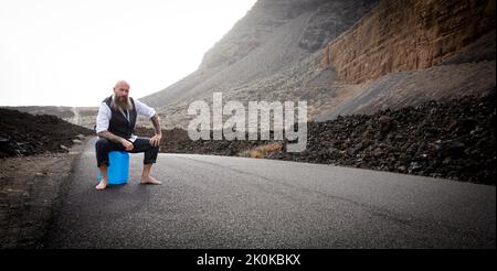 L'uomo con vestito è seduto completamente disperato e bussato fuori su un canister blu da qualche parte in nessun posto nel deserto vulcanico alla fine o Foto Stock