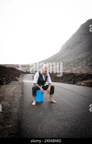 L'uomo con vestito è seduto completamente disperato e bussato fuori su un canister blu da qualche parte in nessun posto nel deserto vulcanico alla fine o Foto Stock