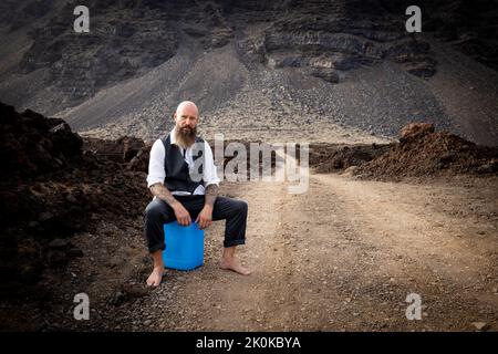 L'uomo con vestito è seduto completamente disperato e bussato fuori su un canister blu da qualche parte in nessun posto nel deserto vulcanico alla fine o Foto Stock