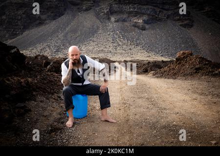 L'uomo con vestito è seduto completamente disperato e bussato fuori su un canister blu da qualche parte in nessun posto nel deserto vulcanico alla fine o Foto Stock