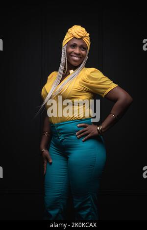 Giovane ragazza afro-americana sorridente con trecce afro tinte che indossano abiti gialli e jeans con foulard e collana su sfondo nero Foto Stock