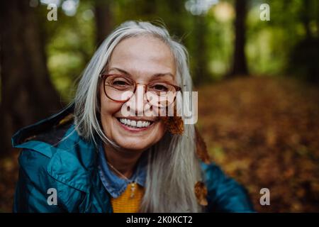 Ritratto di felice donna anziana con foglia sulla guancia a piedi all'aperto nella foresta in autunno. Foto Stock