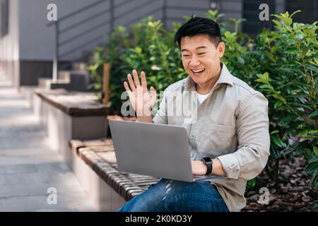 Allegro uomo d'affari asiatico guardando la webcam del computer portatile e la mano di ondulazione, saluto cliente, seduto vicino all'edificio ufficio Foto Stock