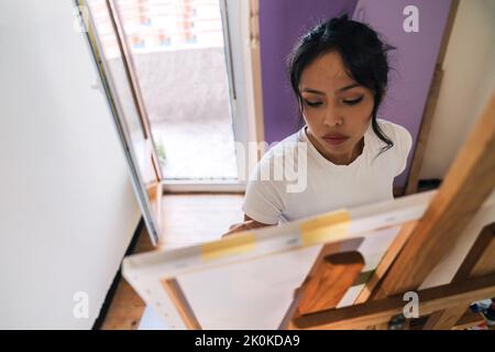 Dall'alto focalizzato etnico femminile in abiti casual pittura su tela con pitture multicolore e pennello in camera piccola e leggera Foto Stock