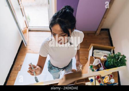 Dall'alto focalizzato etnico femminile in abiti casual pittura su tela con pitture multicolore e pennello in camera piccola e leggera Foto Stock