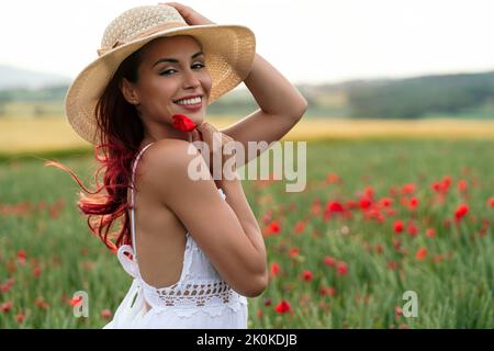 Vista laterale di seducente giovane femmina in cappello di paglia con fiore rosso fiorente sul prato sotto il cielo bianco guardando la macchina fotografica Foto Stock