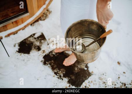 Primo piano di un uomo anziano che tiene un secchio d'acqua ed entra nella sauna all'aperto durante la fredda giornata invernale. Foto Stock