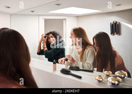 Felice giovane multietnica amici in abbigliamento casual applicare trucco e chattare contro specchio in spogliatoio o bagno Foto Stock