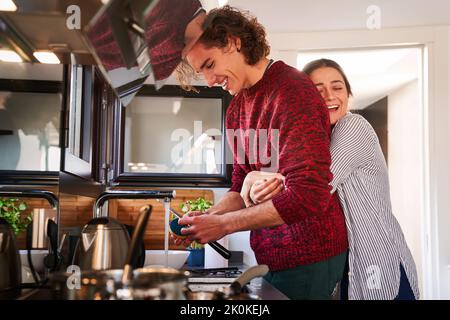 Vista laterale giovane donna deliziata in abiti casual in piedi dietro e abbracciando sorridente ragazzo lavaggio piatti in cucina moderna Foto Stock