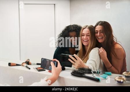 Gruppo di giovani amici multirazziali ridenti che prendono selfie sul telefono cellulare contro lo specchio nello spogliatoio Foto Stock