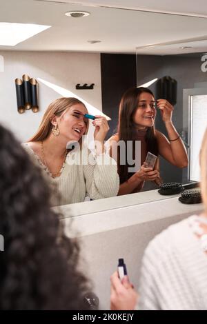 Felice giovane multietnica amici in abbigliamento casual applicare trucco e chattare contro specchio in spogliatoio o bagno Foto Stock