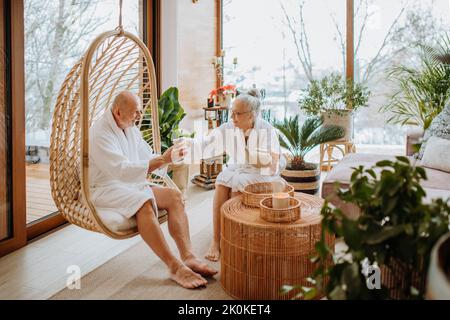 Coppia senior in accappatoi che si gode il tempo insieme nel loro soggiorno, bevendo tè caldo, calma e l'atmosfera hygge. Foto Stock
