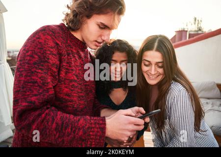 Felice giovani amici multietnici in abiti casual navigazione telefono cellulare, mentre si riuniscono in terrazza in luce del giorno Foto Stock