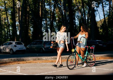 Amici femminili che camminano sulla strada tenendo skateboard e bicicletta in vacanza Foto Stock
