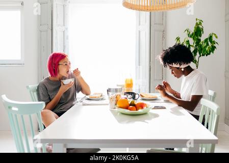 Giovane donna rilassata con i capelli tinti seduta vicino al tavolo e mangiare piatto yummy vicino amico afroamericano in accogliente appartamento Foto Stock