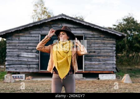 Donna elegante con cappello sul ranch in piedi accanto ad una cabina di legno che guarda lontano Foto Stock