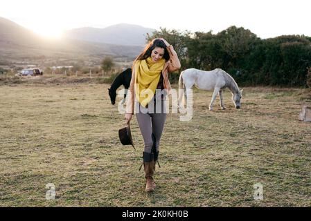 Donna allegra in sciarpa che cammina su campo ranch con cavalli pascolanti alla luce del sole sullo sfondo Foto Stock