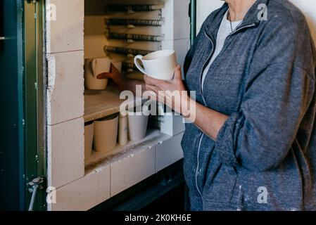 Vista laterale del raccolto irriconoscibile artigiani che collocano tazze di argilla in forno per la cottura di ceramica mentre si lavora in officina Foto Stock