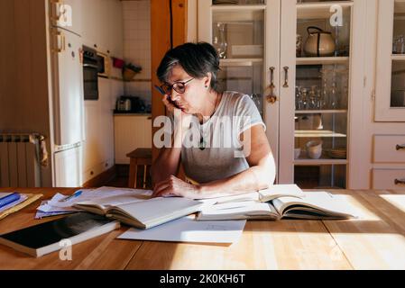 Donna di mezza età messa a fuoco in occhiali con penna in mano seduta a tavolo con un mucchio di libri e carte e studiare materiali professionali a casa Foto Stock