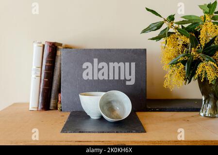 Composizione di vita morta con ciotole bianche in ceramica poste su tavola di ardesia su tavola di legno con vecchi libri e vaso con fiori fioriti Foto Stock