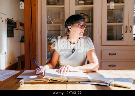 Donna di mezza età messa a fuoco in occhiali con penna in mano seduta a tavolo con un mucchio di libri e carte e studiare materiali professionali a casa Foto Stock