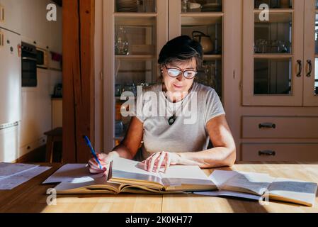 Donna di mezza età messa a fuoco in occhiali con penna in mano seduta a tavolo con un mucchio di libri e carte e studiare materiali professionali a casa Foto Stock