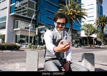 Sorridente imprenditore maschile bearded in abbigliamento formale e occhiali da sole che hanno video chat su smartphone mentre si siede sulla panca in strada soleggiata vicino al bui moderno Foto Stock