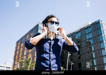 Da sotto di maschio in maschera medica e occhiali da sole ascoltando musica attraverso le cuffie, mentre in piedi in strada soleggiata vicino a edifici moderni durante la co Foto Stock