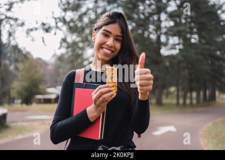 Soddisfare giovane studentessa ispanica con libri che mostrano pollice in su gesto mentre godendo sano barretta di cereali durante la pausa nel parco verde nel campus Foto Stock