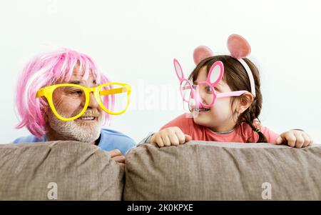 Bambino eccitato in finti occhiali con coniglietto e fascia con orecchie mouse con nonno in grandi finti occhiali e parrucca rosa che si guardano l'un l'altro whi Foto Stock