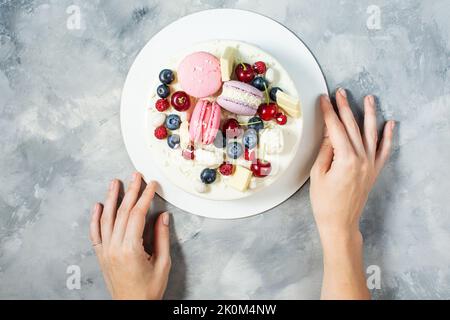 Dolce torta bianca decorata con cioccolato bianco fuso, amaretti, meringhe, bacche e caramelle su fondo in cemento. Foto Stock