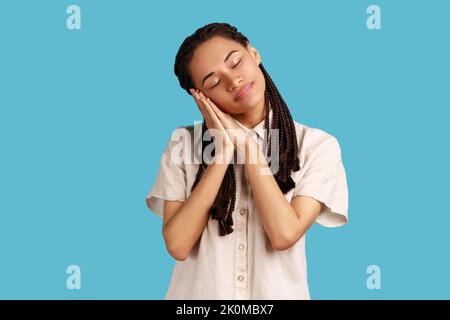 Ritratto di bella donna con dreadlocks neri che dorme adagiandosi sulle sue palme, avendo comodo pelo e riposo, spianando fuori, indossando una camicia bianca. Studio in interni isolato su sfondo blu Foto Stock