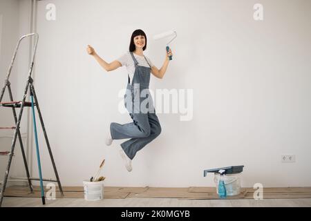 Felice donna gioiosa, giovane ragazza caucasica capelli rossi saltando mentre dipinge il muro con rullo di vernice bianco. Riparazione nel concetto di appartamento. Foto Stock