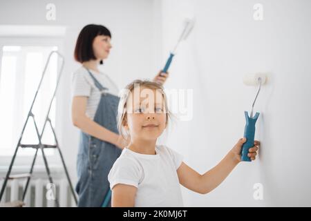 Sorridente ragazza di 4 anni tiene un rullo di vernice nelle sue mani. Il bambino aiuta sua madre, che sta dietro di lei, vicino al muro, nella riparazione. Foto Stock