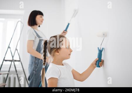 Sorridente ragazza di 4 anni tiene un rullo di vernice nelle sue mani. Il bambino aiuta sua madre, che sta dietro di lei, vicino al muro, nella riparazione. Foto Stock