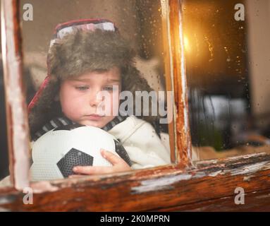 HES ha ottenuto il blues piovoso di giorno. Un bambino triste che tiene una sfera di calcio mentre osserva la pioggia attraverso una finestra. Foto Stock