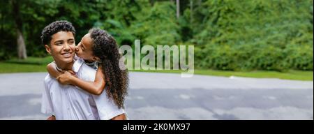 Una sorellina sulla schiena del suo grande fratello adolescente dandogli un bacio sulla guancia in un momento accattivante tra fratelli in famiglia Foto Stock