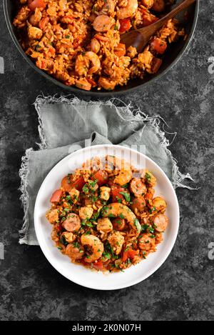 Jambalaya creola con gamberi, salsicce affumicate, carne di pollo e verdure sul piatto su fondo di pietra scura. Vista dall'alto, disposizione piatta Foto Stock