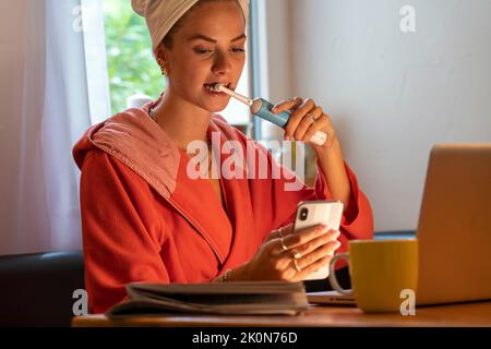 Immagine simbolica Precrastinazione, giovane donna cerca di fare molte cose allo stesso tempo, dopo il bagno, sul computer, telefono, giornale, e colazione Foto Stock